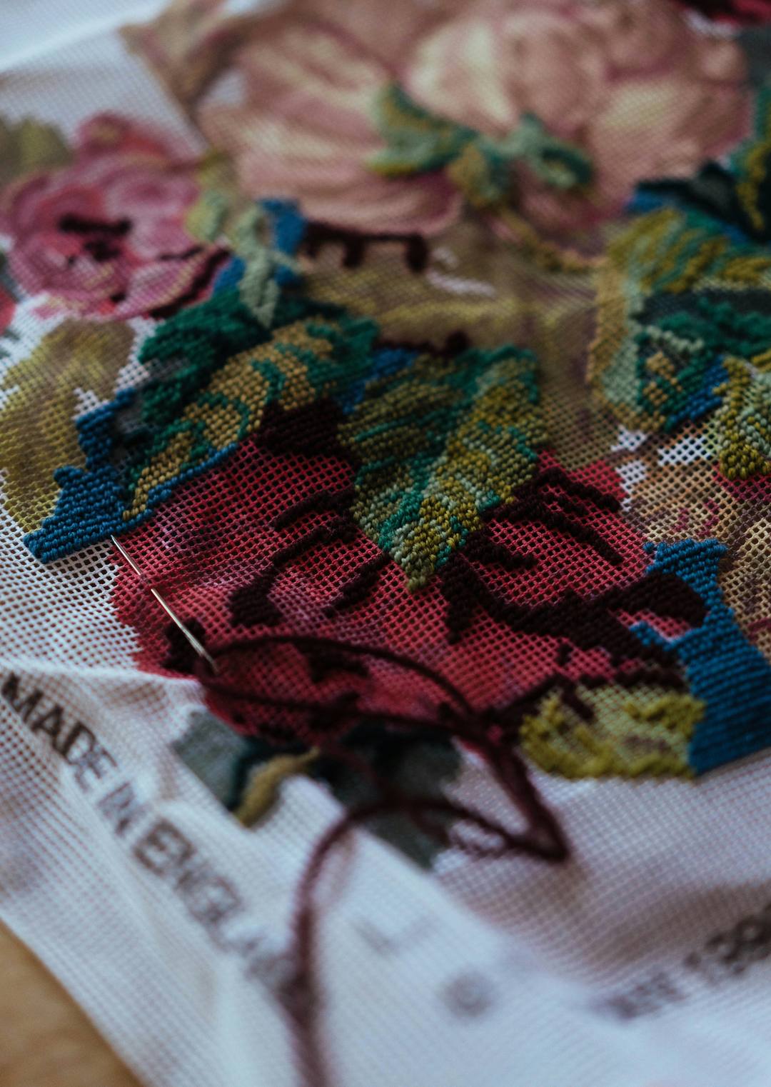 A needle mid-stitch on a floral needlepoint canvas, with deep red, teal, and moss green wool half worked.