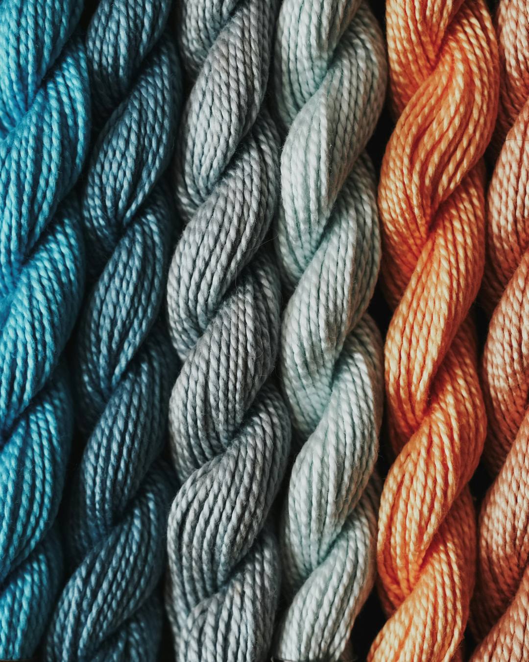 Twisted skeins of needlepoint thread in a row, shading from deep blue through gray to orange and rust.